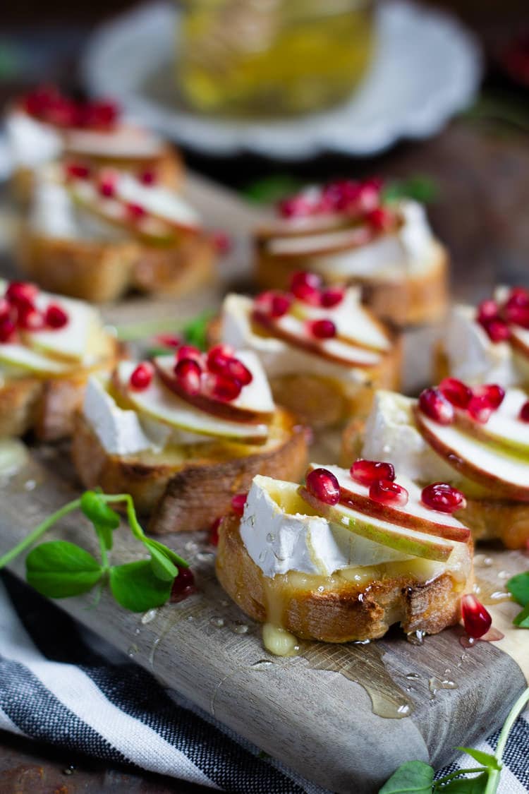 A wooden cutting board serving platter loaded with apple brie crostini garnished with honey and pomegranate seeds