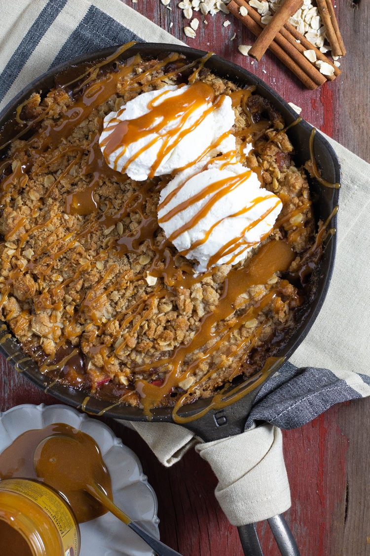 Cast iron pan filled with homemade apple crisp drizzled with caramel and topped with vanilla ice cream