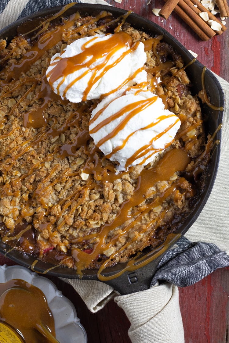 Cast iron pan filled with homemade apple crisp drizzled with caramel and topped with vanilla ice cream