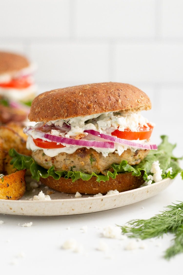 Straight on photo of a Greek turkey burger on a wheat bun topped with lettuce, feta, tomato, red onion and tzatziki