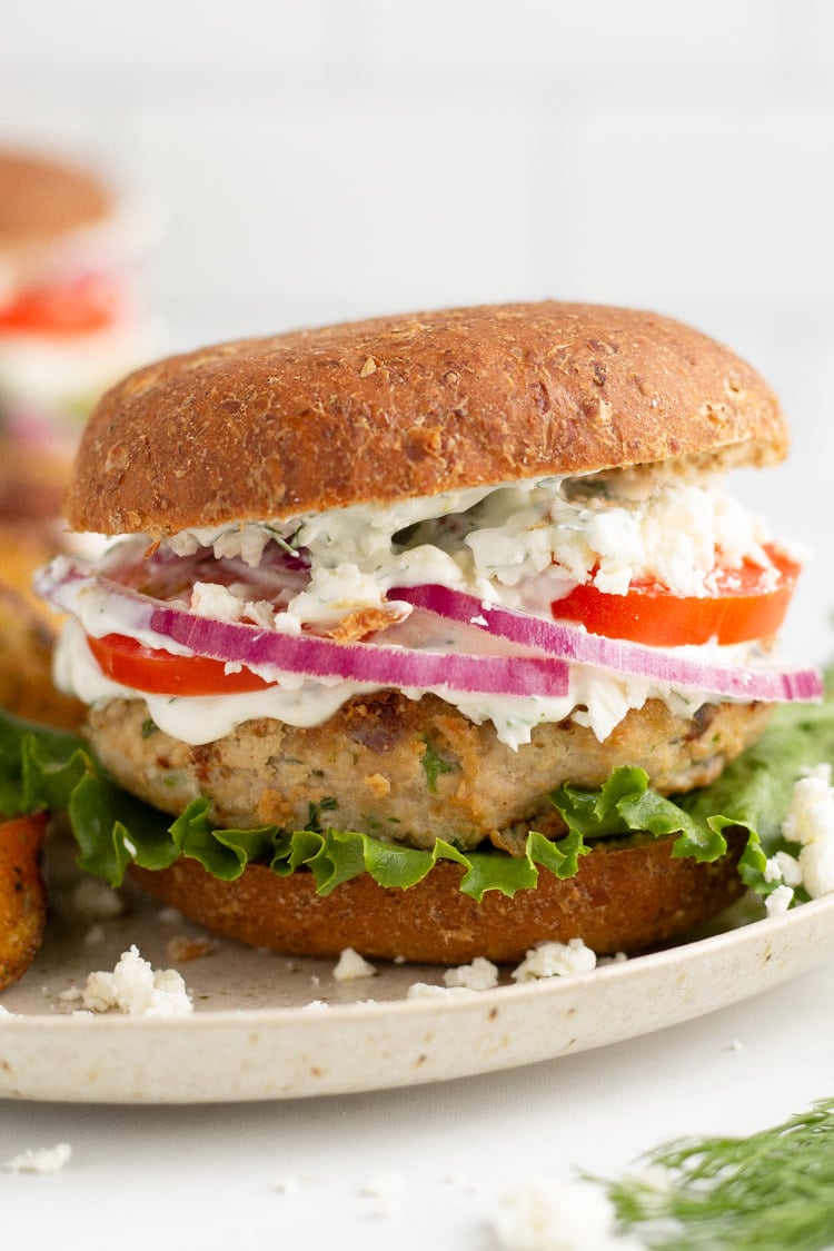 Straight on photo of a Greek turkey burger on a wheat bun topped with lettuce, feta, tomato, red onion and tzatziki