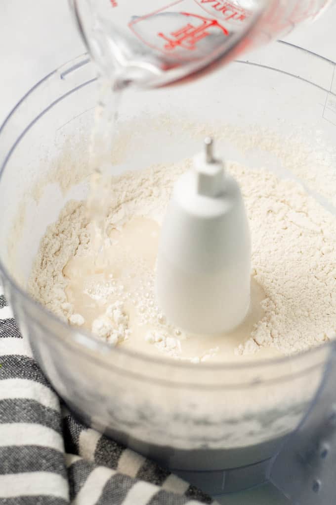 Close up shot of a food processor with flour and a measuring cup of water being poured in