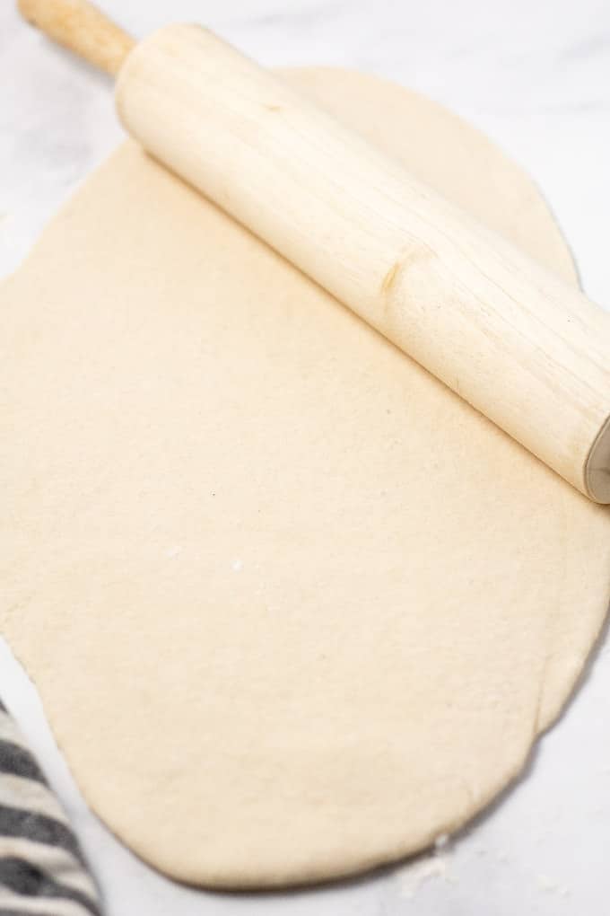 A rolling pin with a large oval of flatbread pizza dough rolled out on a white marble counter top