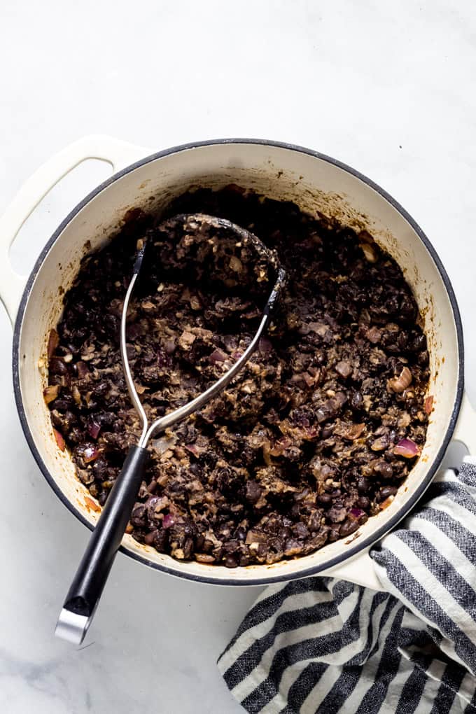 Overhead shot of a white pot with a potato masher mashing black beans and onions