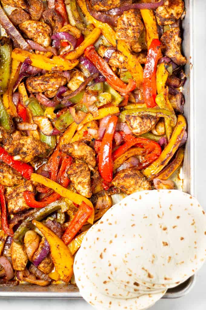 Overhead shot of a metal sheet pan loaded with peppers onions and chickens with flour tortillas.