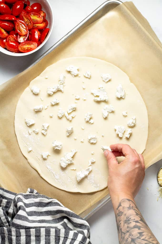 Pie crust on a baking sheet being topped with crumbled goat cheese