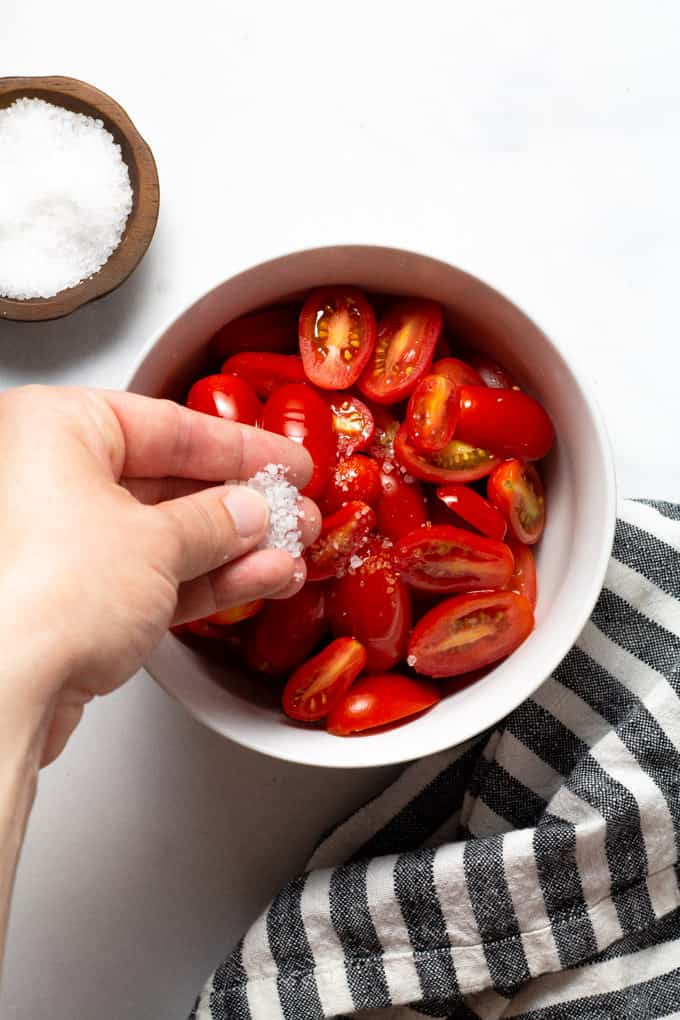White bowl of tomatoes being sprinkled with Kosher salt