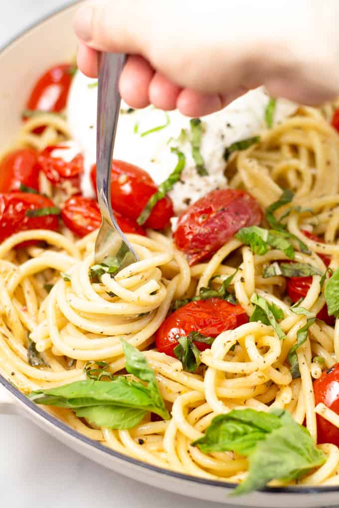 Close up shot of a fork twirling pasta in a pan filled with tomato and garlic pasta garnished with fresh basil
