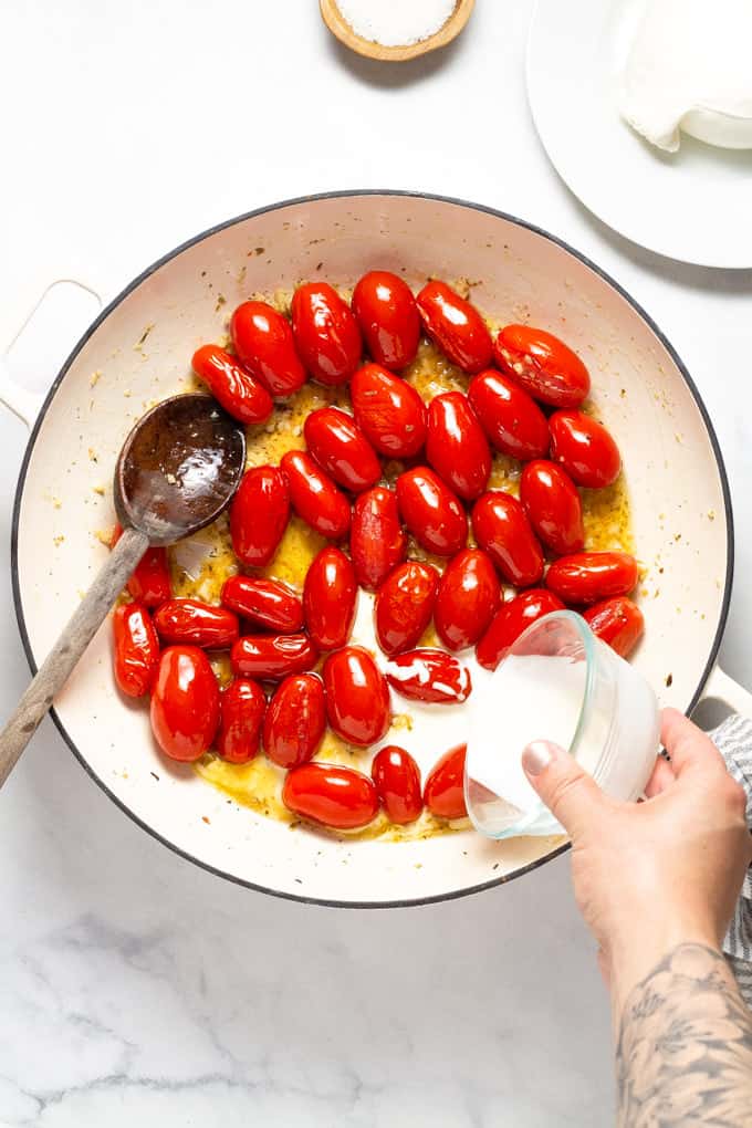 Overhead shot of a pan filled with burst cherry tomatoes and cream being poured into the pan