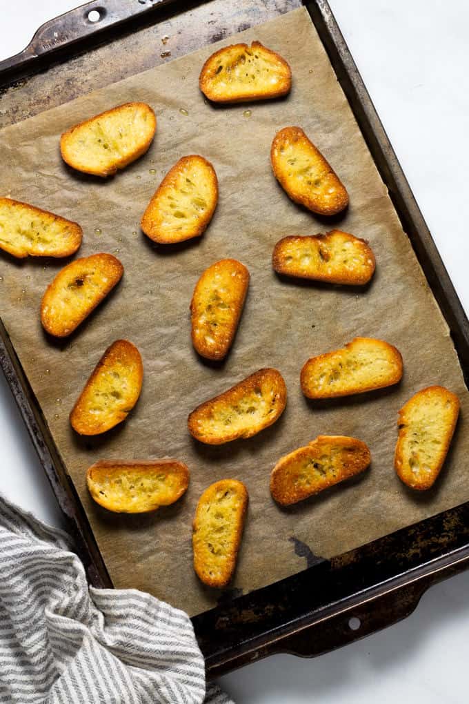 Slices of French baguette on a parchment lined baking sheet after being broiled in the oven 