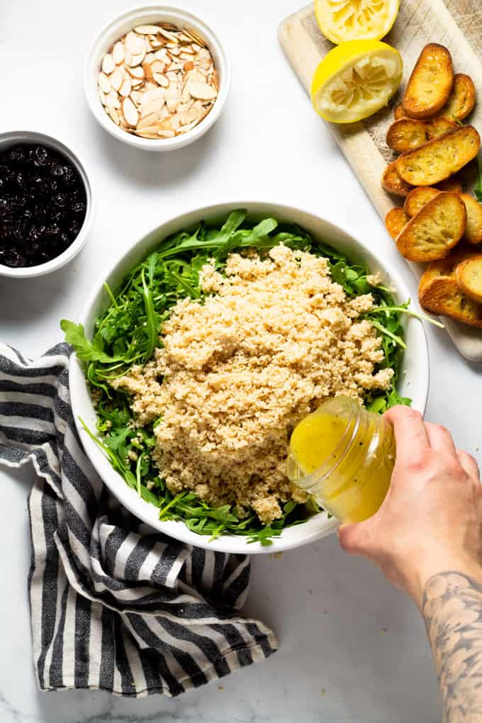 A white bowl filled with arugula and couscous with lemon vinaigrette being poured over it 