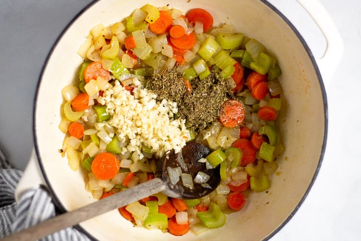 White dutch oven with cooked carrots onion celery garlic and herbs in it