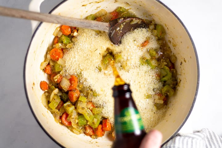 White dutch oven with cooked veggies and beer being poured into it