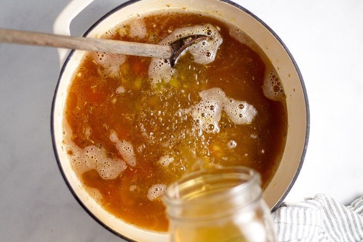 White dutch oven with cooked veggies and chicken broth being poured into it