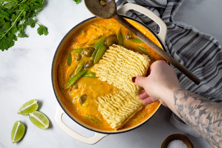 Overhead shot of a white dutch oven filled with curry vegetables with ramen noodles being added