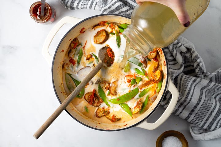 Overhead shot of a white dutch oven filled with curried vegetables with vegetable broth being added