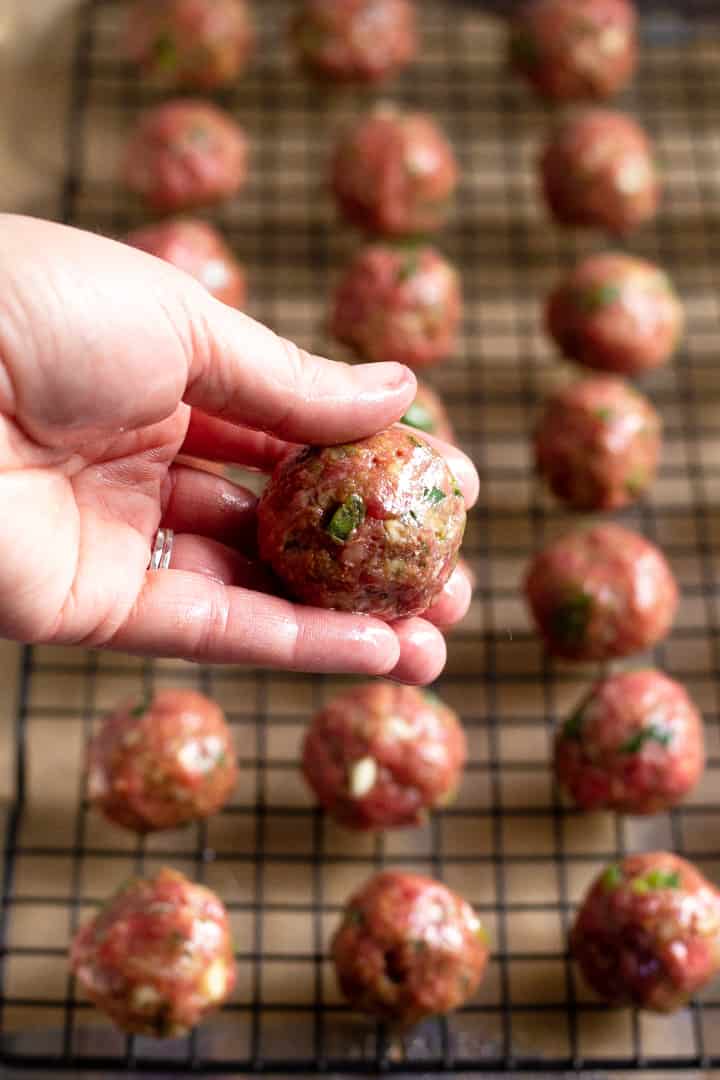 A close up shot of a raw meatball with a baking sheet of meatballs in the background 