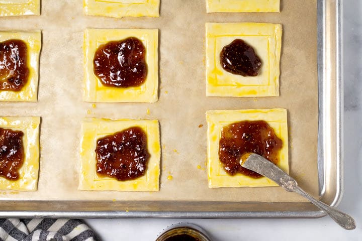 Puff pastry squares on a parchment lined baking sheet being spread with fig jam 