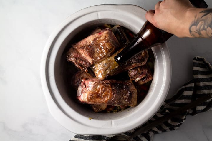 Large crock pot insert filled with seared short ribs with beer being poured over them