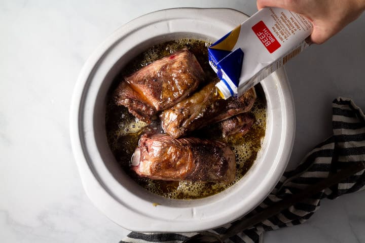 Large crock pot insert filled with seared short ribs with beer broth being poured over them