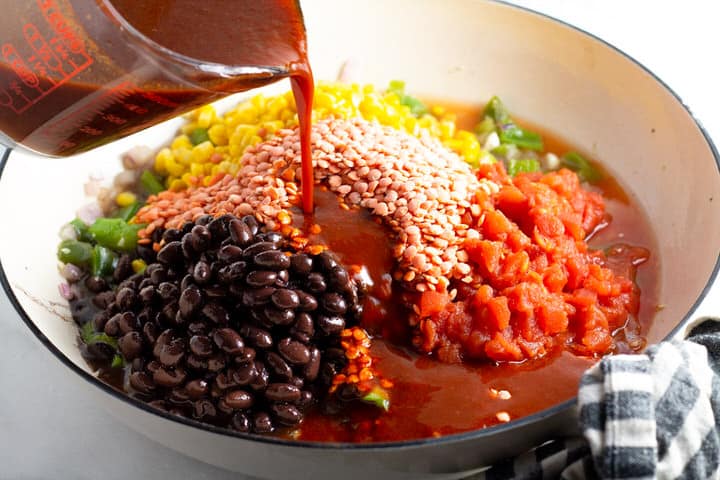 White saute pan filled with veggies with a cup of enchilada sauce being poured over it