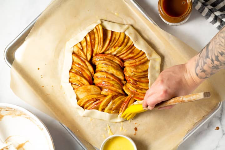 Pie crust filled with sliced apples coated in sugar and spices being brushed with egg wash