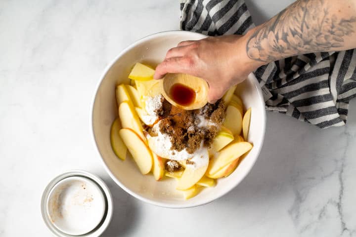 White bowl of sliced apples and spices with vanilla extract being poured in