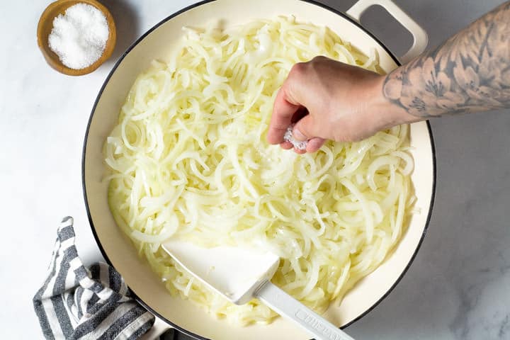 Overhead shot of a white pan filled with thinly sliced onions with salt being added to them