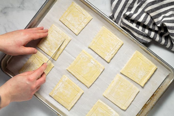 Puff pastry squares on a parchment lined baking sheet with a knife scoring the crust of each square