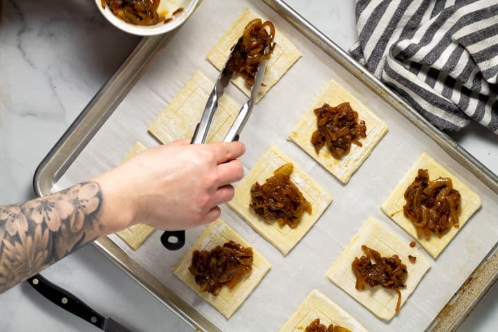 Caramelized onions being add to puff pastry squares on a parchment paper lined baking sheet