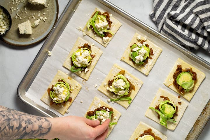 Blue cheese being added to puff pastry squares on a baking sheet