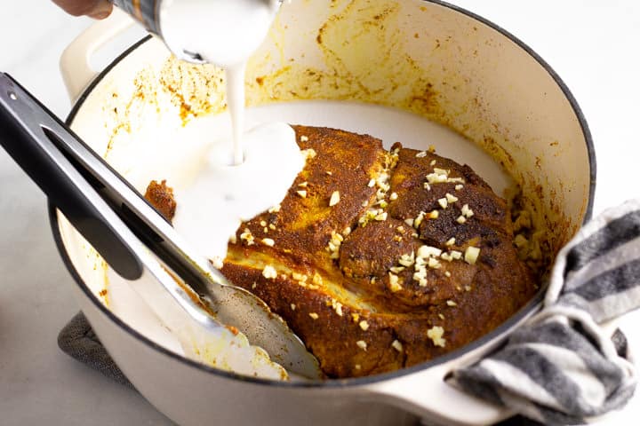 Large white pot filled with seared pork roast with coconut milk being poured over it