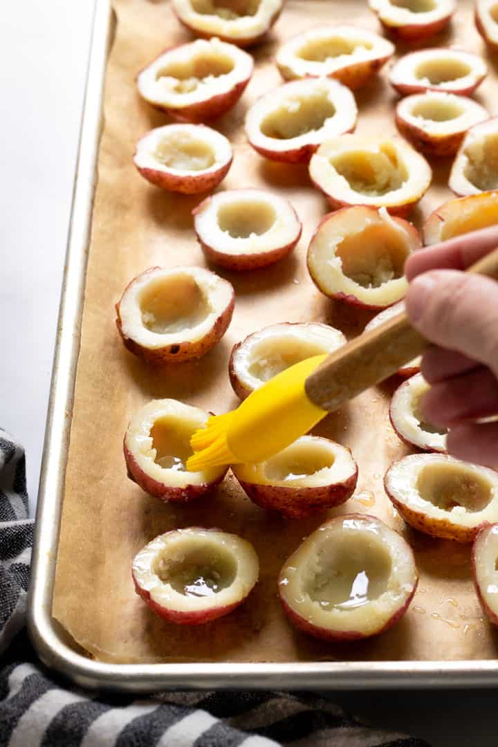 A parchment lined baking sheet with halved baked potatoes with the flesh scooped out of the center being brushed with oil 
