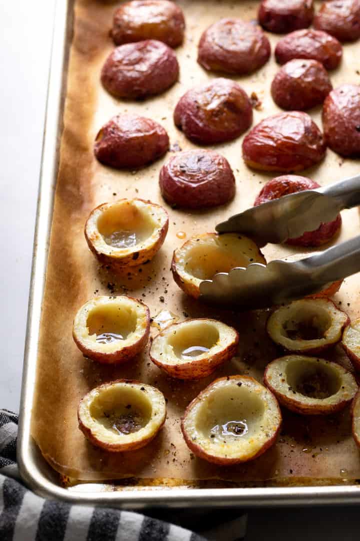 A parchment lined baking sheet with halved baked potatoes with the flesh scooped out of the center being flipped with tongs