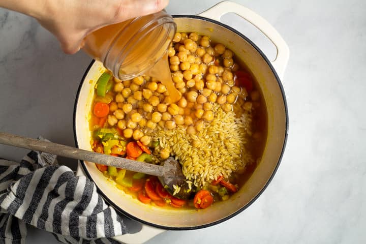 Large white dutch oven filled with vegetables chickpeas and orzo pasta with broth being poured in