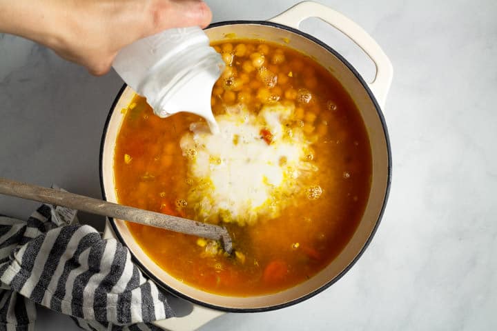 Large white dutch oven filled with vegetables and chickpeas with coconut milk being poured in
