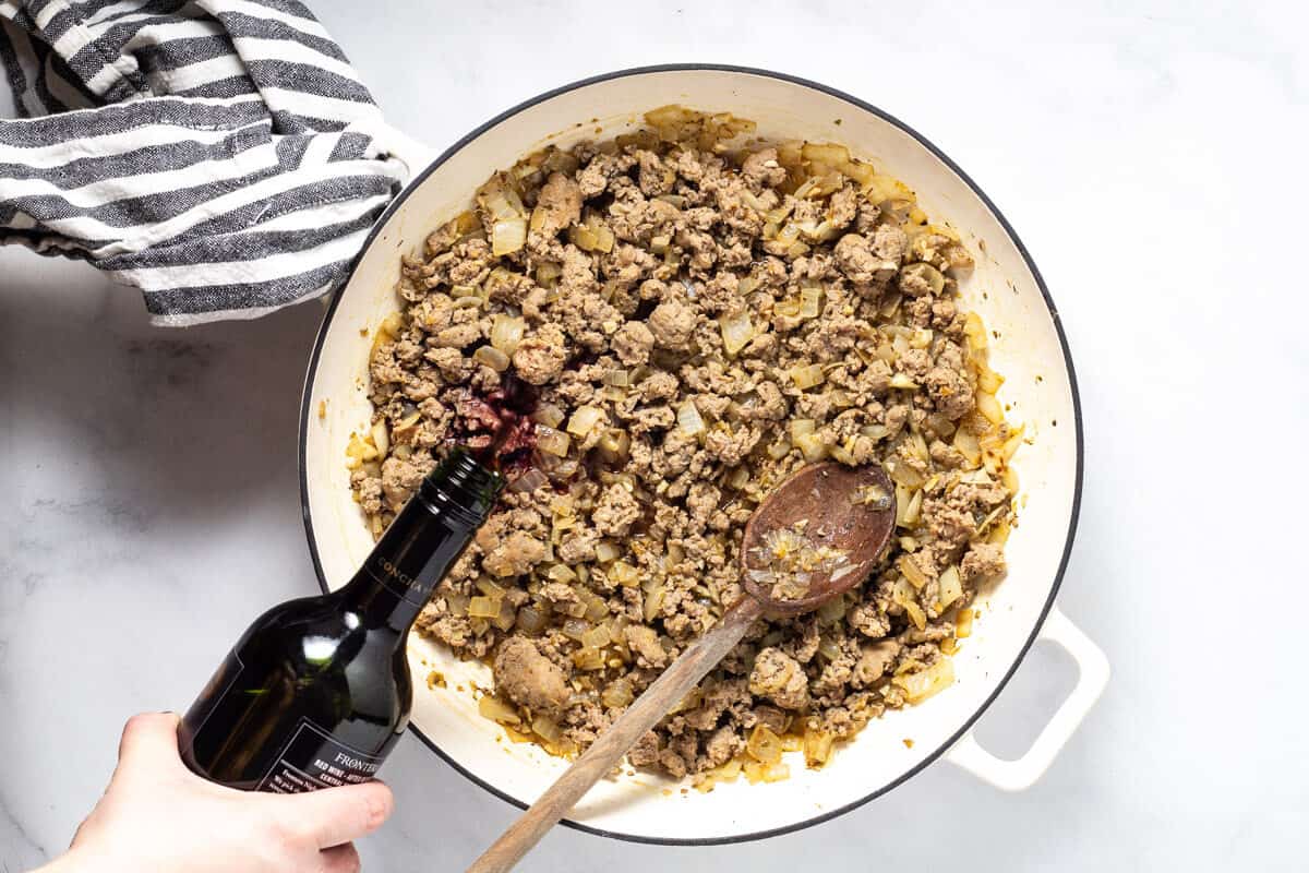 Large white saute pan filled with browned sausage with red wine being poured in
