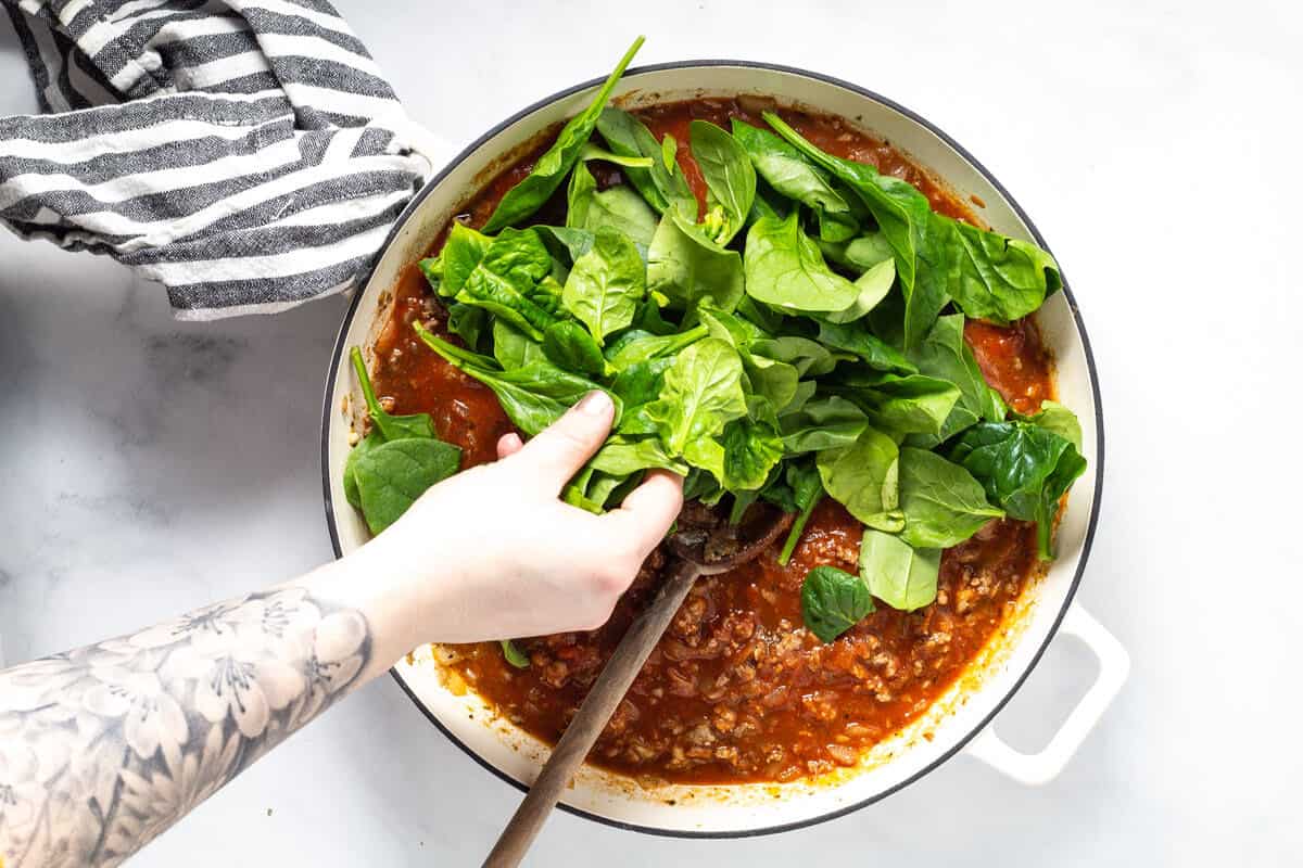 Large white saute pan filled with spaghetti sauce with fresh spinach being added to it