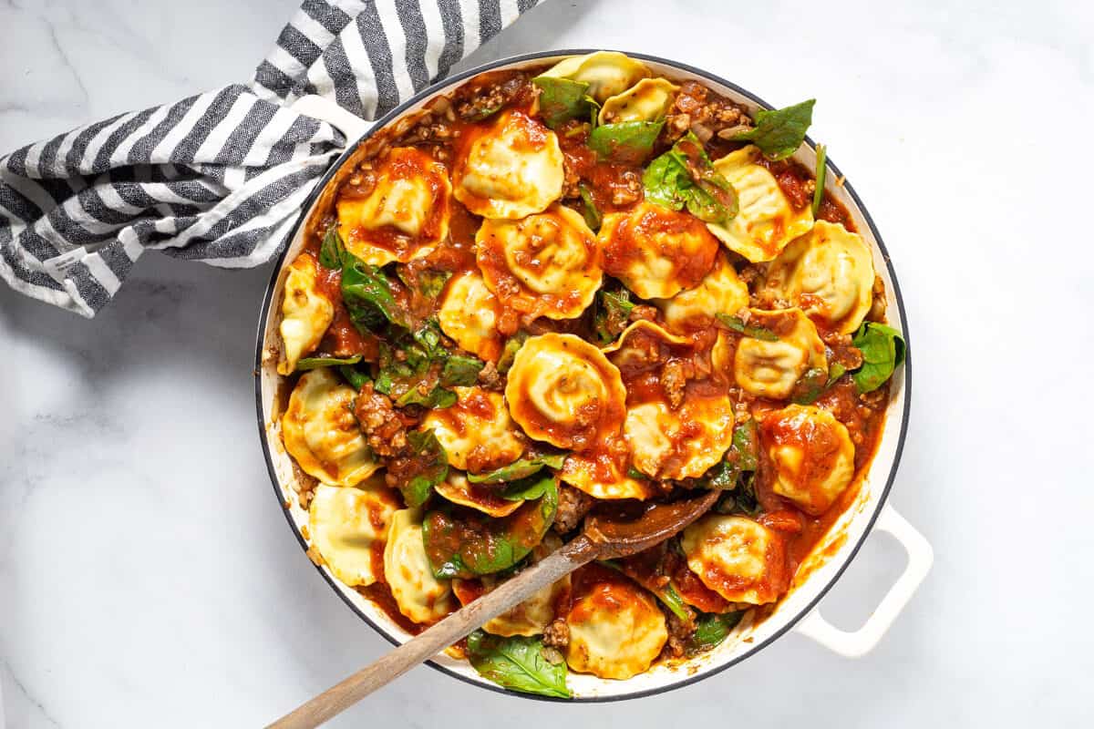 Large white saute pan filled with spaghetti sauce spinach and round ravioli