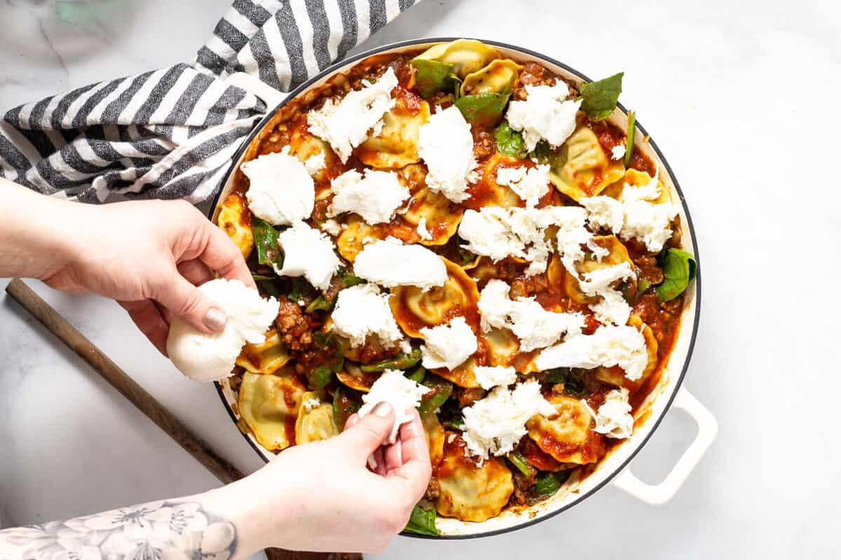 Large white saute pan filled with spaghetti sauce spinach ravioli and fresh mozzarella being added
