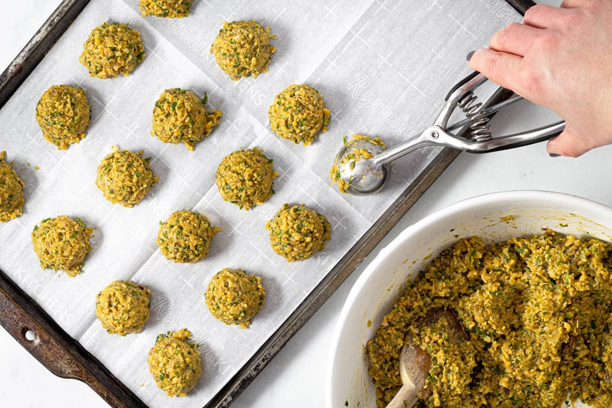 A cookie scoop scooping balls of falafel onto a parchment lined baking sheet