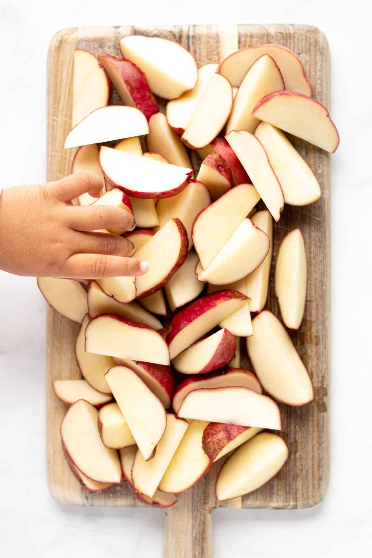 Overhead of a wooden cutting board with sliced potato wedges and a little hand reaching in and grabbing a wedge
