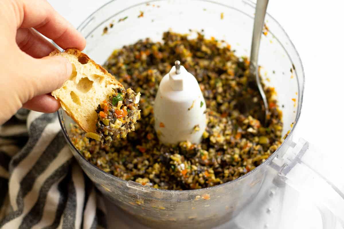 Hand using a slice of bread to scoop olive tapenade out of a food processor
