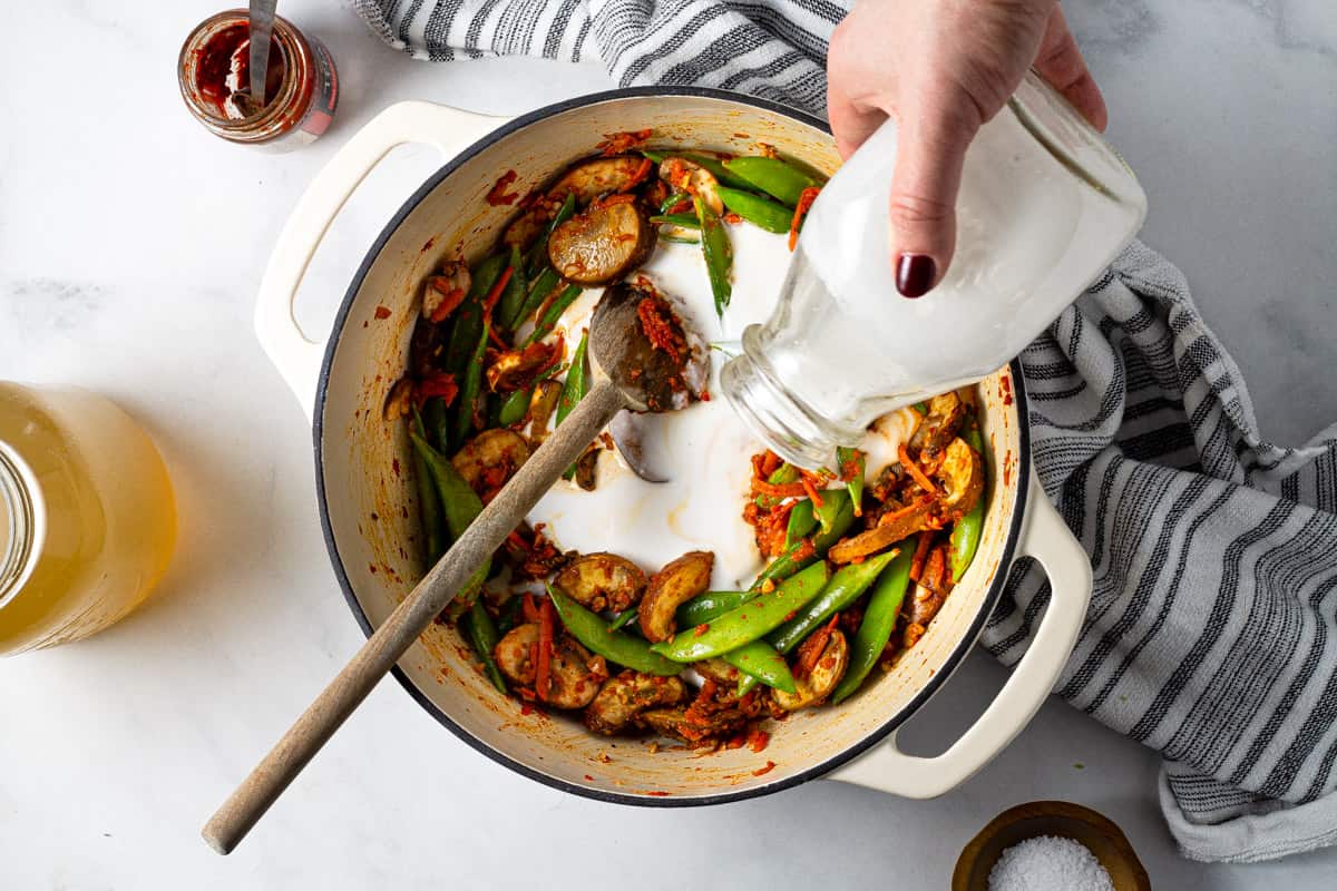 Overhead shot of a white dutch oven filled with curried veggies with coconut milk being added.