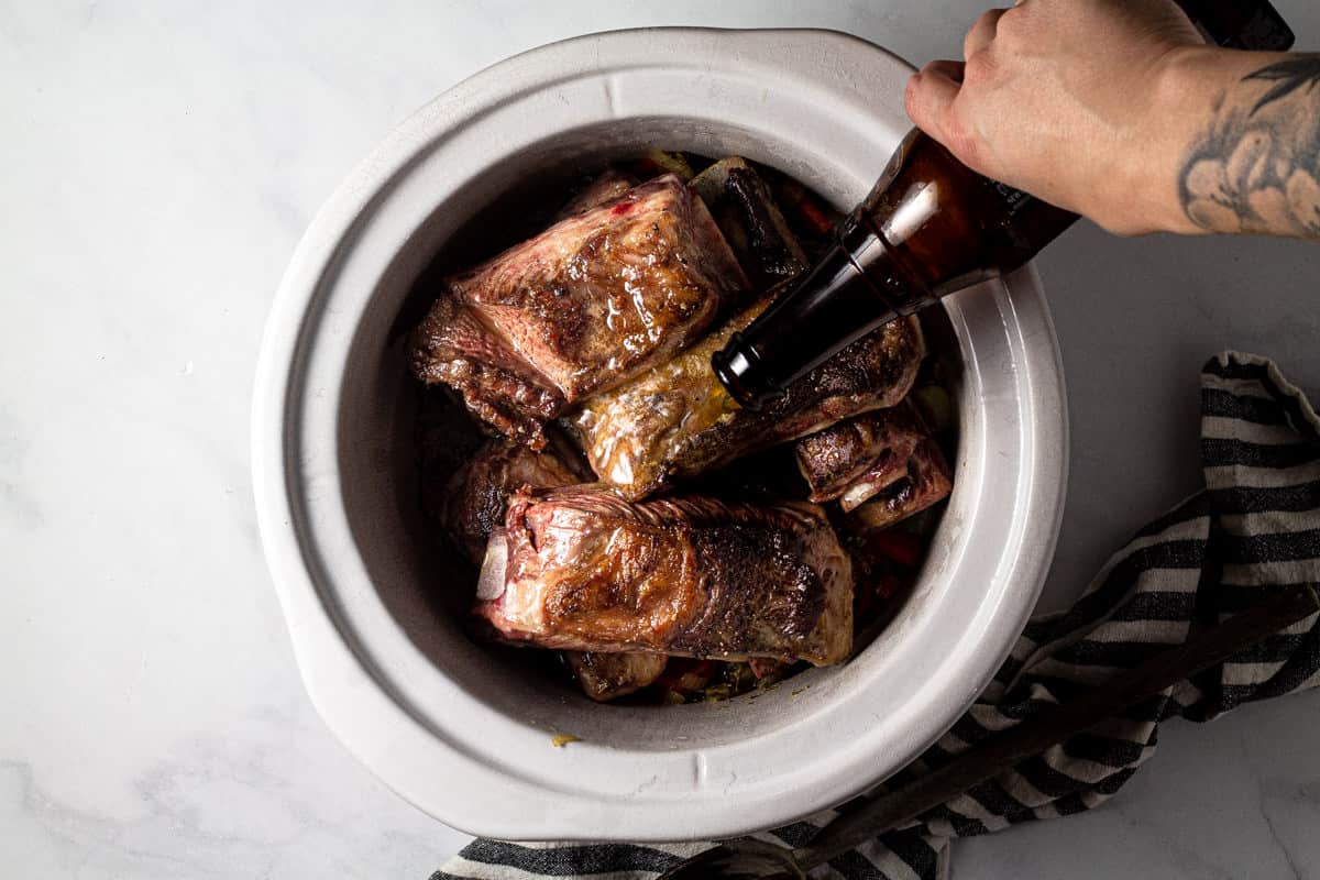 Large crock pot insert filled with seared short ribs with beer being poured over them