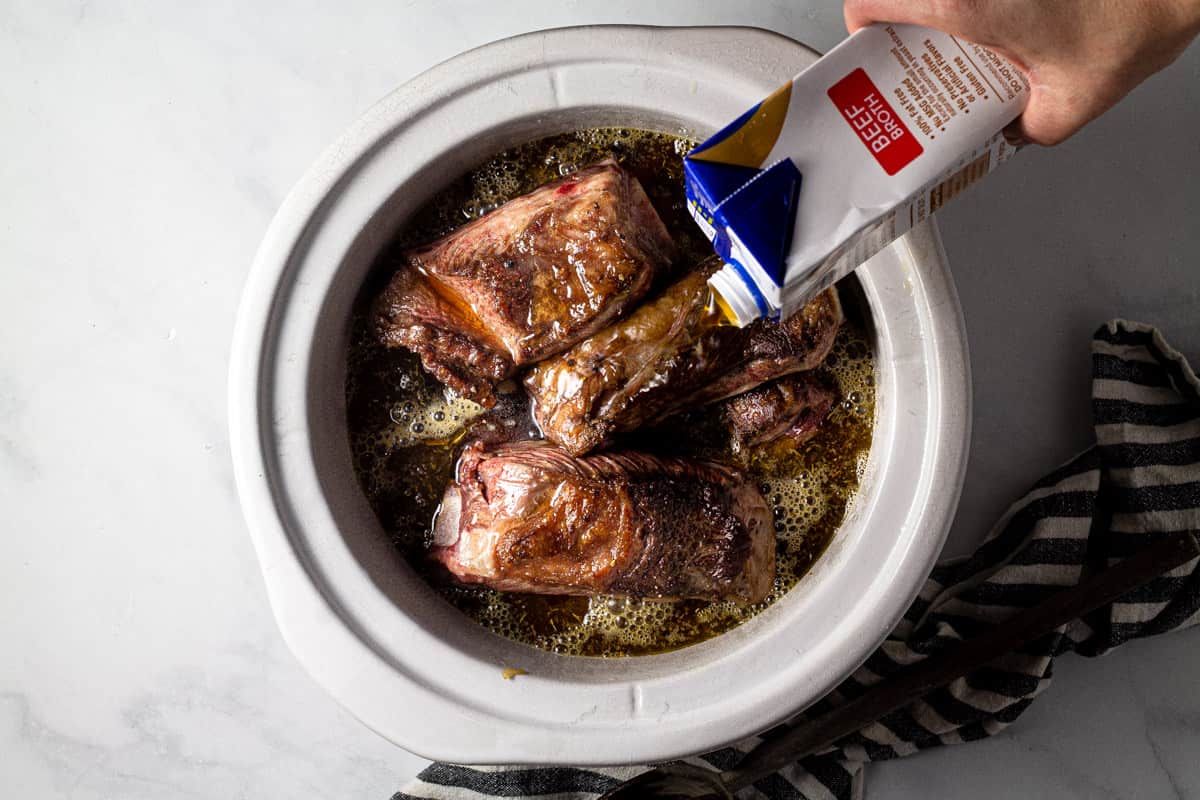 Large crock pot insert filled with seared short ribs with beer broth being poured over them 
