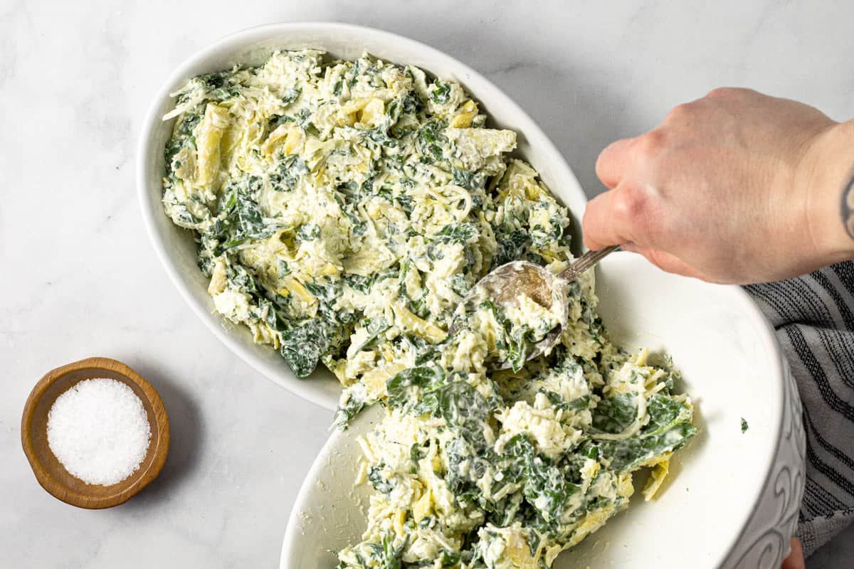 Spinach dip being transferred from a white bowl to a white baking dish