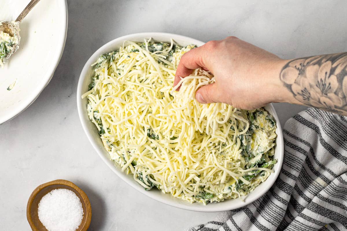 Spinach dip in a white baking dish being topped with shredded cheese