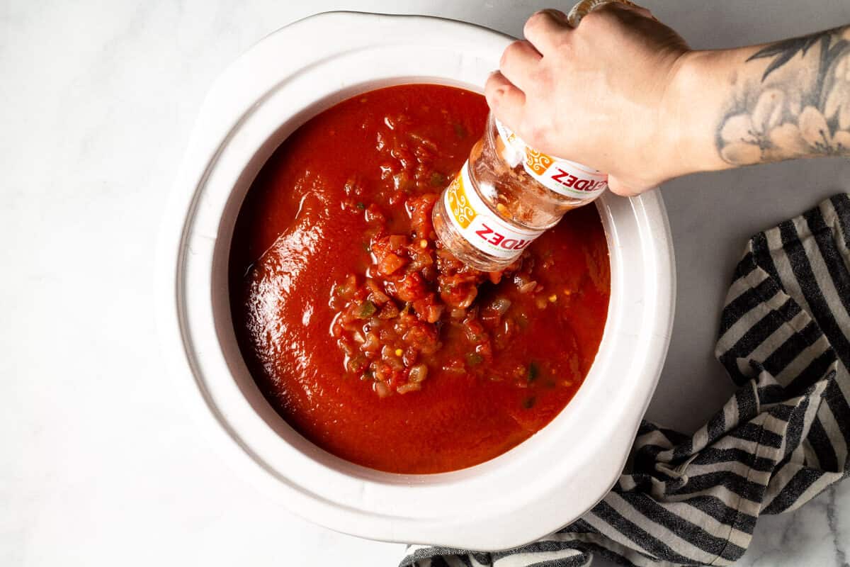 A hand pouring a jar of salsa into a white crock pot insert