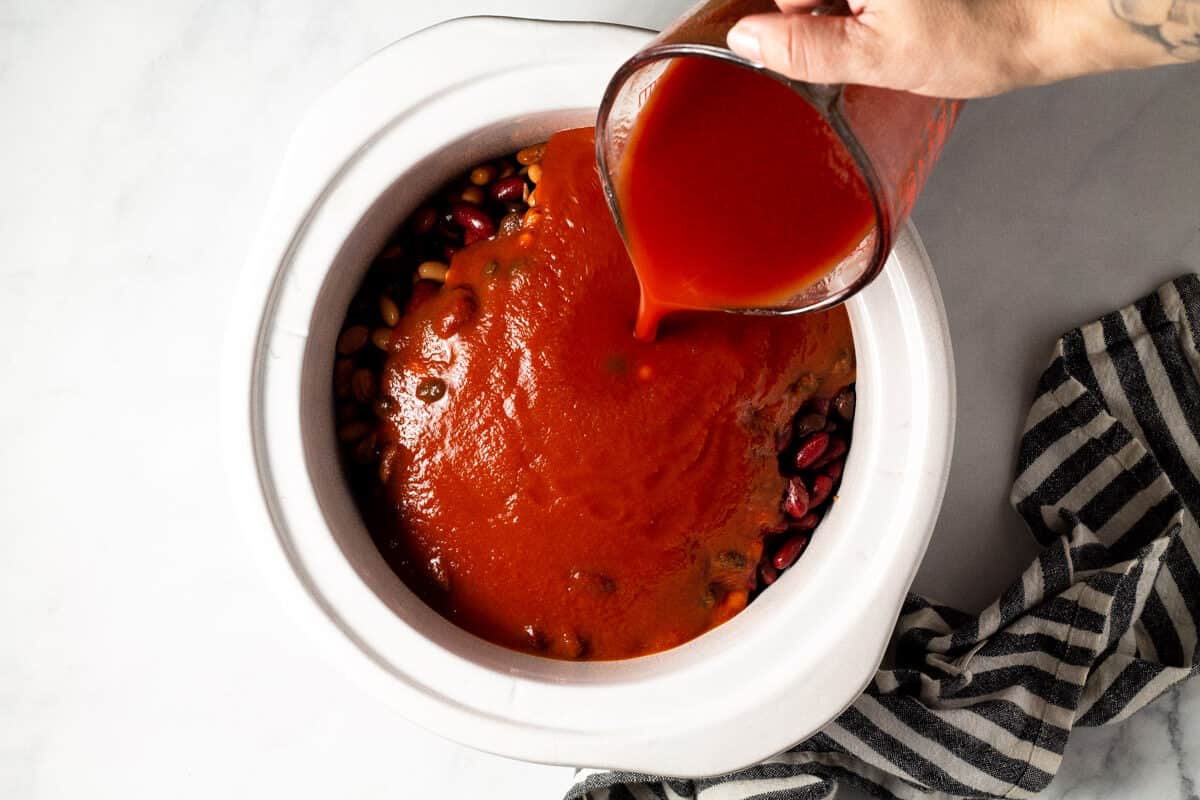 A hand pouring tomato sauce into a white crock pot insert